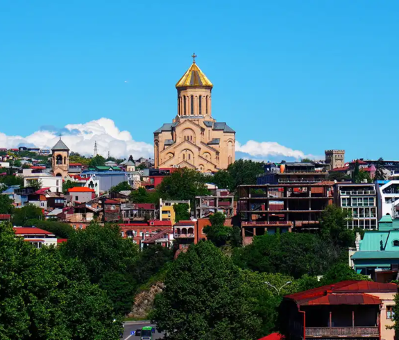 A panoramic view of Holy Trinity Cathedral of Tbilisi rising above green trees and colorful residential buildings under a bright blue sky, ideal for budgeting Georgia Travel Cost.