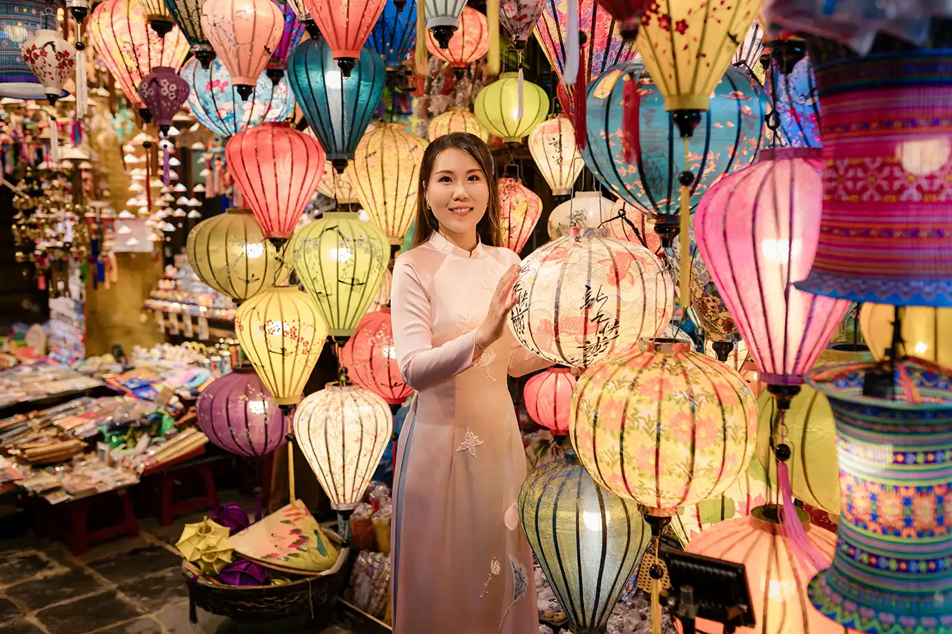 A medium portrait photograph of a smiling Vietnamese woman wearing a traditional light pink Ao Dai dress, gently touching a round lantern while surrounded by colorful, illuminated silk lanterns at Hoi An Night Market in Vietnam.