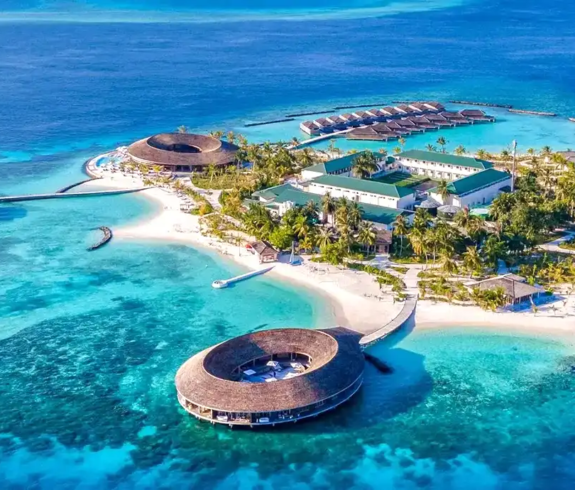 A high-angle panoramic aerial photograph looking down on the secluded island of Kagi Maldives Resort & Spa, featuring a distinct circular overwater thatched-roof spa, pristine white sand beaches, overwater villa jetties, and turquoise lagoon waters surrounded by the deep blue ocean.
