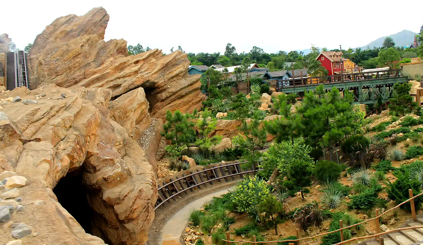 A roller coaster track winding through rugged red rock formations, caves, and Western-themed wilderness in Grizzly Gulch, featured in a Hong Kong Disneyland Guide.