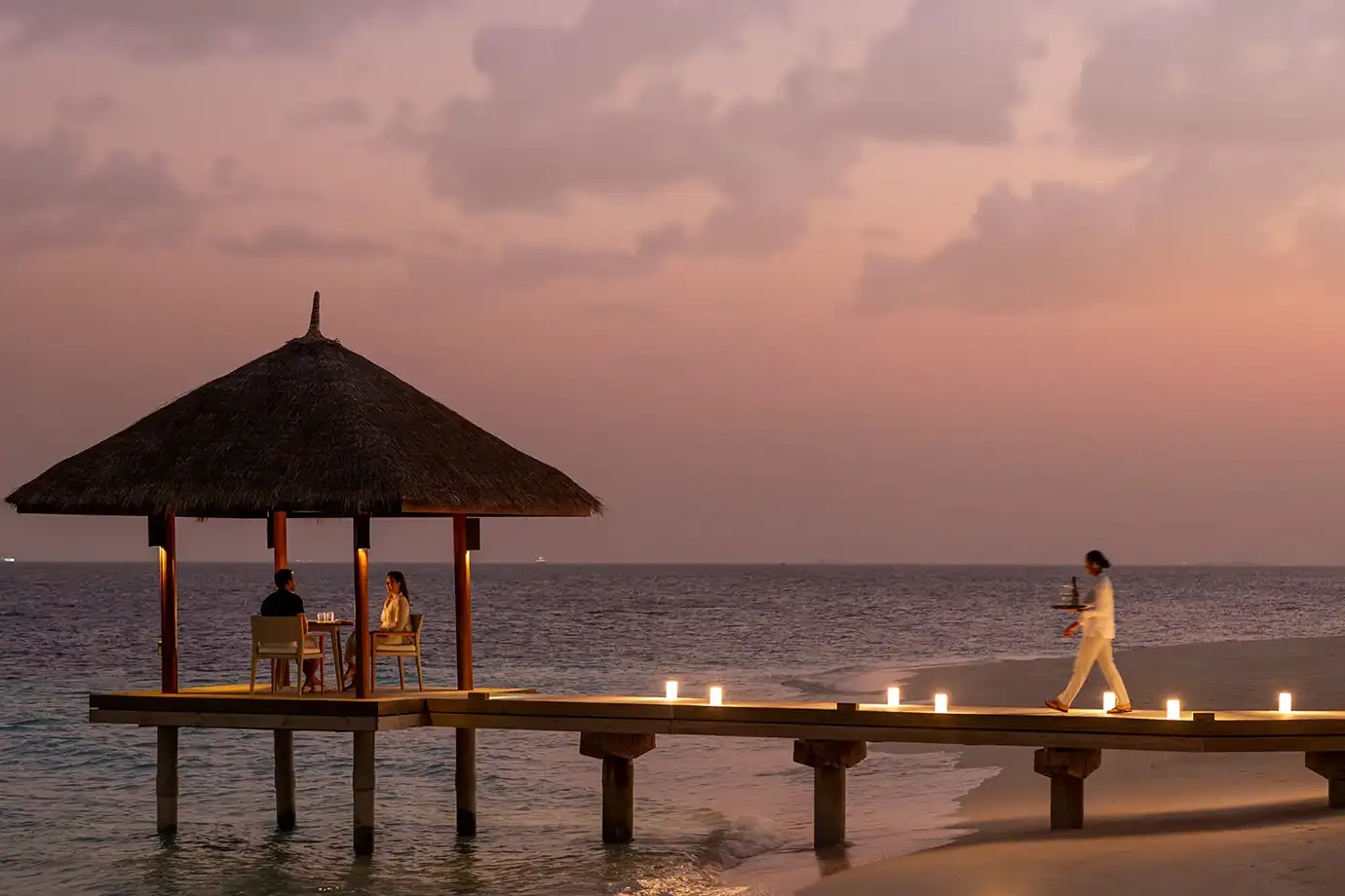 A wide, eye-level dusk photograph showing a couple dining inside an illuminated thatched-roof wooden overwater pavilion while a waiter approaches along a jetty lit with soft lantern lights, set against a dusky pink and purple sky in the Maldives.