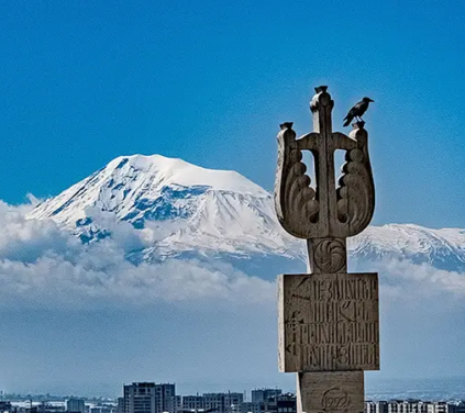 Best of Armenia Tour - A vertical telephoto photograph showing a detailed carved stone monument topped with a bird in the foreground, overlooking the city skyline of Yerevan and the snow-capped peak of Mount Ararat under a deep blue sky in Armenia.