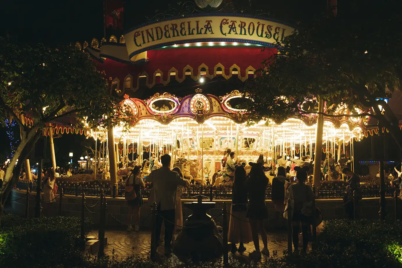 The brightly illuminated Cinderella Carousel glowing at night with guests watching from the courtyard, featured in a Hong Kong Disneyland Guide.