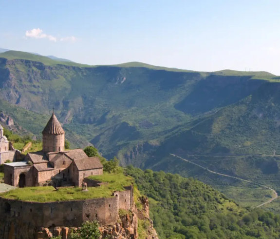 A wide landscape photograph of the fortified Tatev Monastery complex situated on a steep cliff edge overlooking the expansive green valleys and rolling mountains of the Vorotan River Gorge under a clear sky in Armenia.