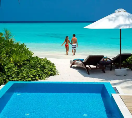 Maldives Honeymoon Tour - A high-angle landscape photograph of a private resort villa terrace with a bright blue tiled plunge pool, wooden sun loungers under a white umbrella, and a couple in swimwear walking toward vibrant turquoise ocean waters on a white sand beach in the Maldives.