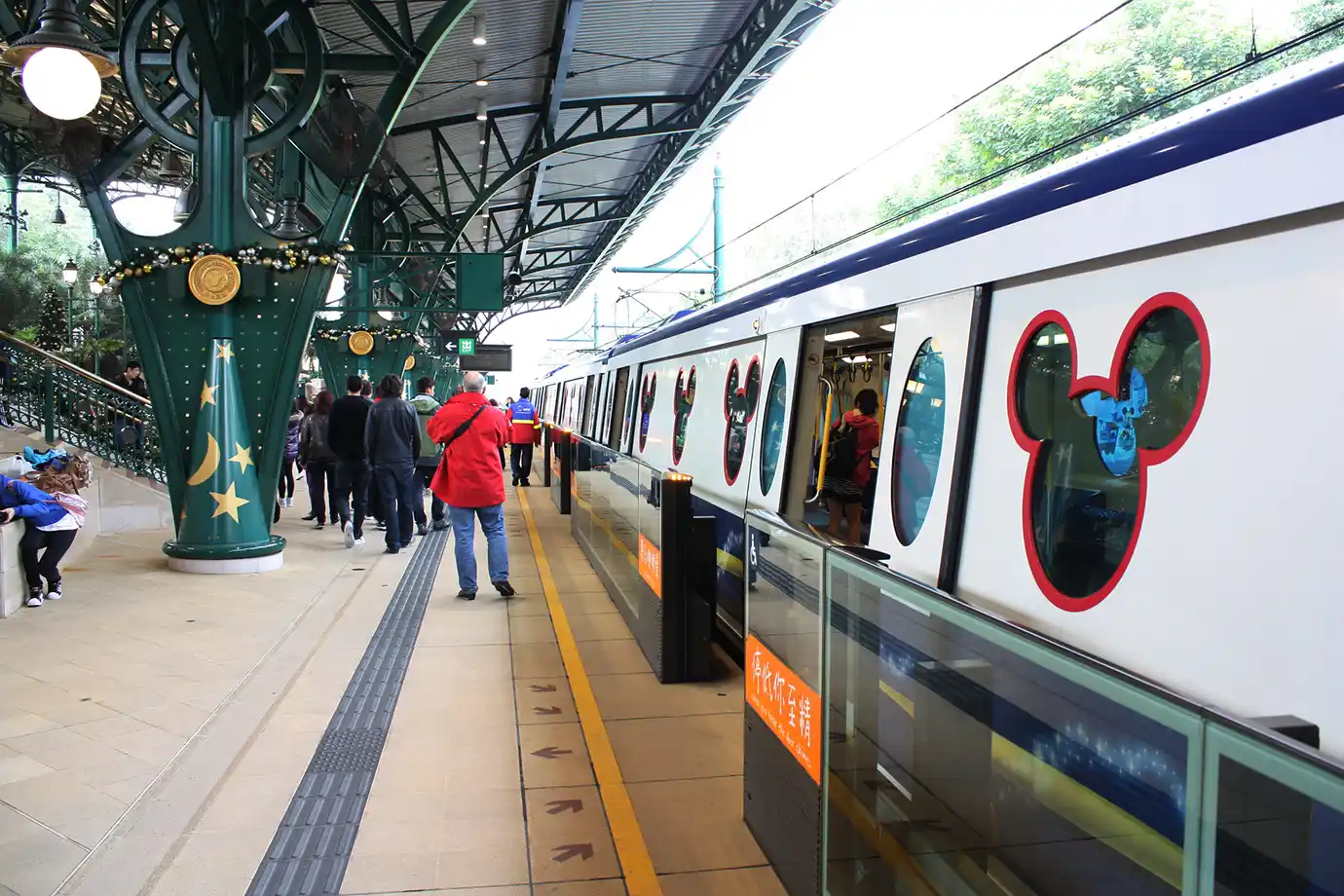 A themed MTR train with Mickey Mouse-shaped windows stopped at a covered station platform with passengers boarding, featured in a Hong Kong Disneyland Guide.