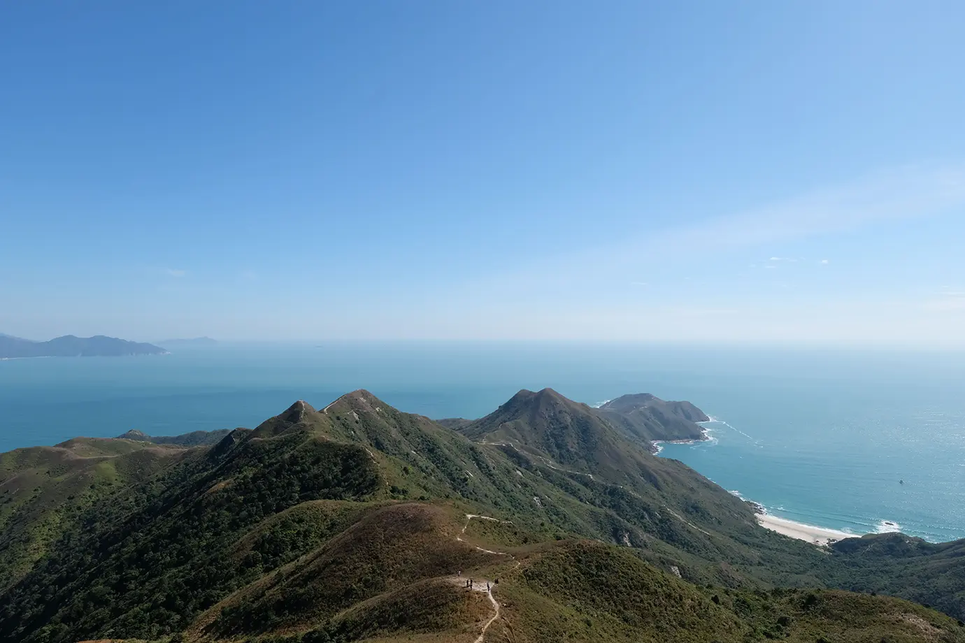 A high-angle scenic photograph of hikers walking along the undulating ridge of Dragon's Back trail with panoramic views of the ocean, coastline, and sandy beach below, highlighting outdoor Things to Do in Hong Kong. 