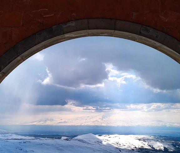A symmetrical, eye-level architectural photograph looking outward through a dark red tuff-stone stone archway covered in snow, framing a distant view of snow-capped Mount Ararat and rolling highlands beneath dramatic sunlit clouds in Yerevan, Armenia.