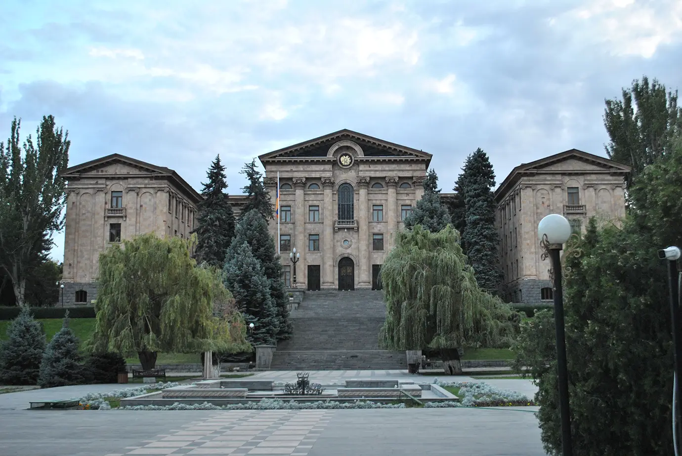 A wide, eye-level exterior photograph showing the pink tuff-stone National Assembly of Armenia building with a central grand staircase, Corinthian columns, and manicured green trees under an overcast sky in Yerevan.
