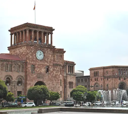 Yerevan Tour - A wide outdoor photograph of the pink and yellow tuff stone Neoclassical Government House featuring a prominent clock tower with an Armenian flag, set beside singing fountains and leafy trees in Republic Square, Yerevan, Armenia.