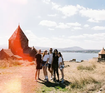Armenia Grand Tour - A sunny film-style photograph showing a group of five friends posing together on a grassy hill in front of the stone Sevanavank Monastery churches with Lake Sevan and distant mountains in the background under a scattered cloud sky in Armenia.