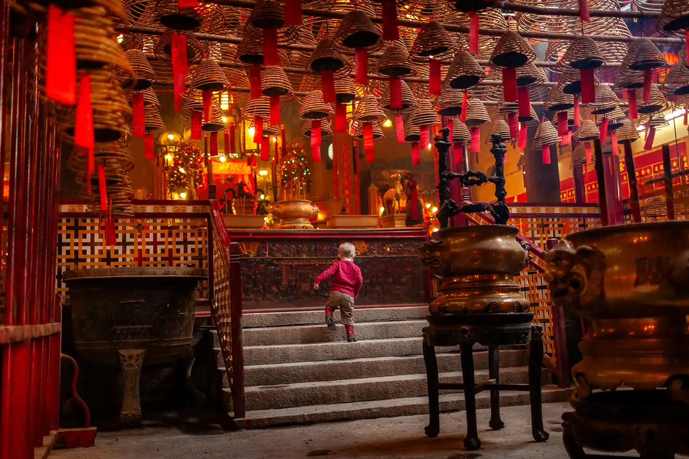 A child walking up steps inside an atmospheric historic temple beneath numerous burning incense coils hanging from the ceiling, representing cultural experiences for Things to Do in Hong Kong. 