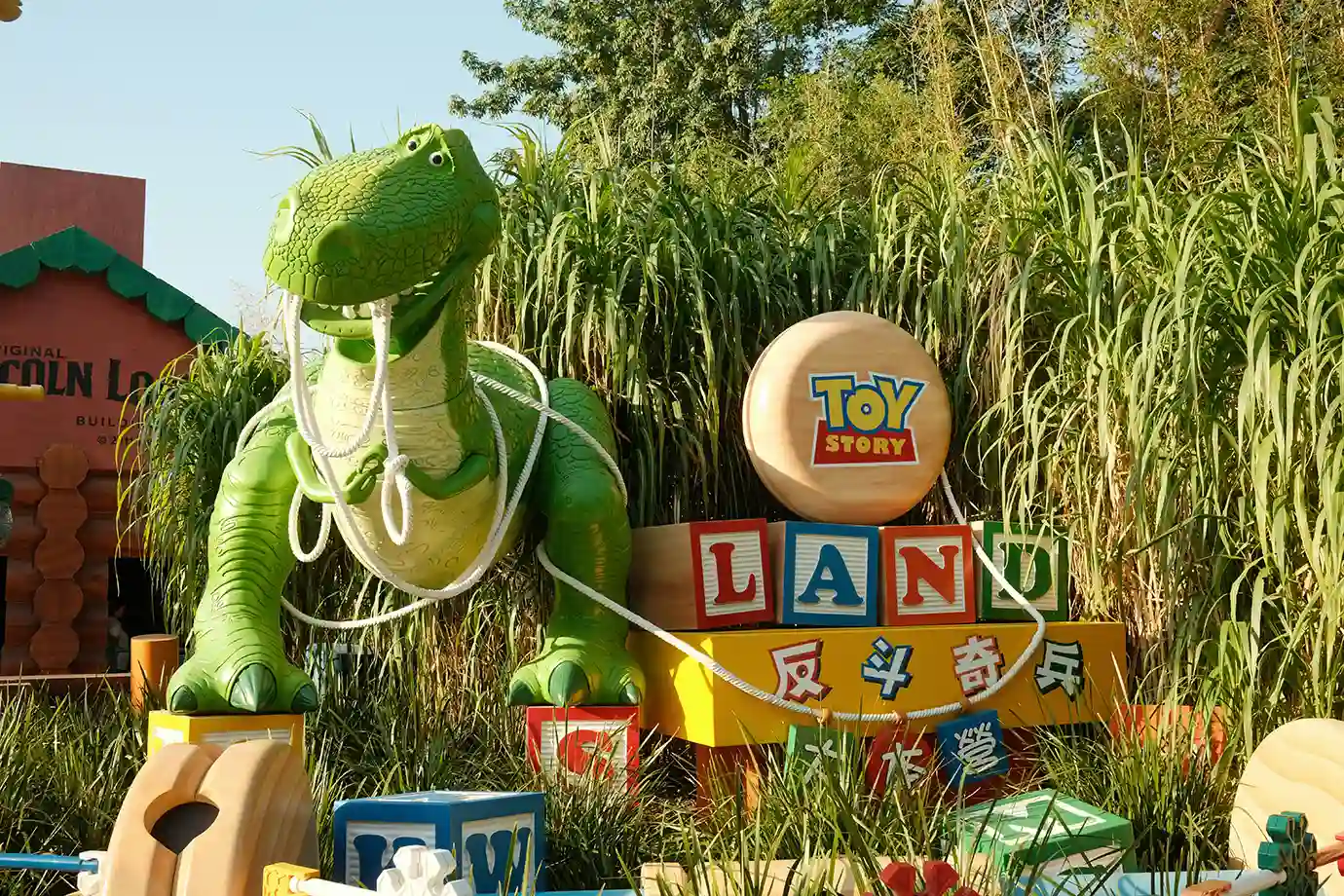 A large green Rex the dinosaur toy with rope and colorful alphabet blocks spelling out the Toy Story Land entrance sign, featured in a Hong Kong Disneyland Guide.