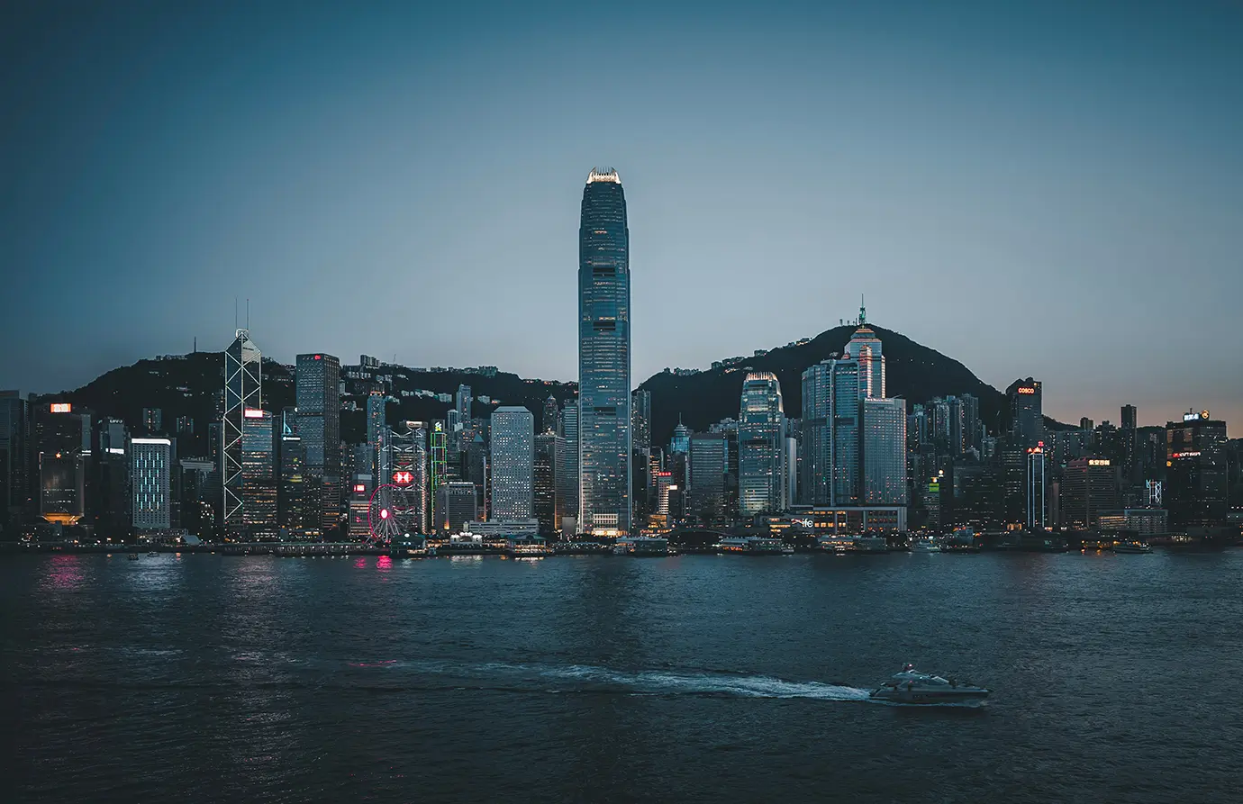 A wide dusk view of the illuminated Hong Kong skyline across Victoria Harbour with iconic high-rises and a boat gliding on the water, highlighting top activities for Things to Do in Hong Kong. 