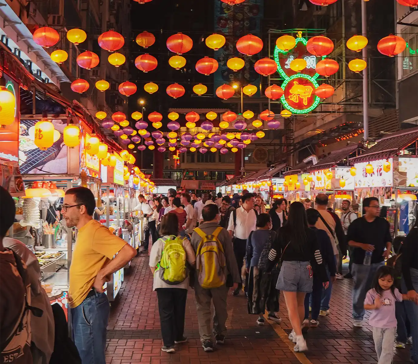 A bustling night market street filled with visitors strolling past brightly lit food stalls beneath rows of hanging red and yellow paper lanterns, highlighting evening activities for Things to Do in Hong Kong. 