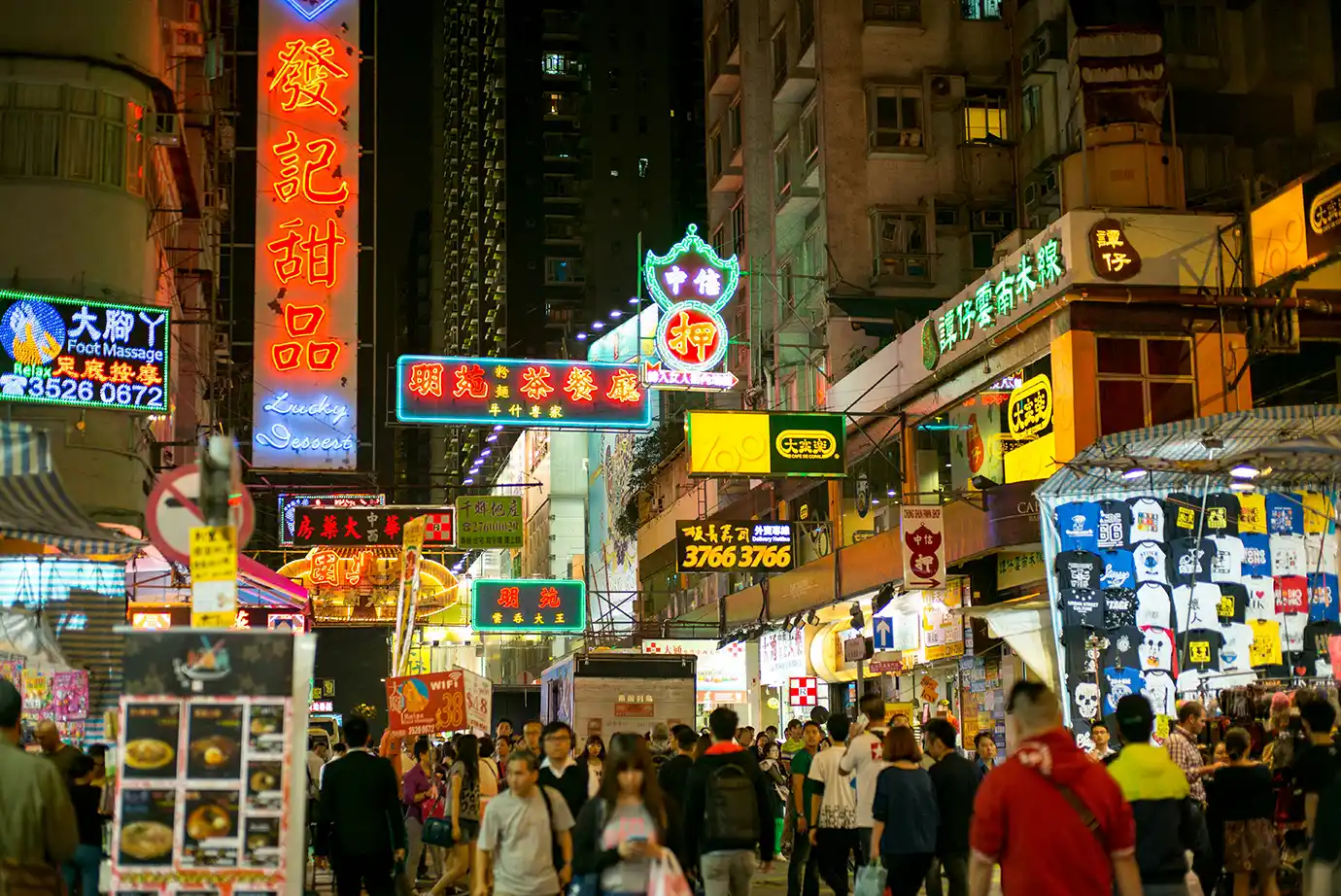 A bustling nighttime street scene in Mong Kok filled with crowds of shoppers walking beneath glowing neon signage and market stalls, representing top urban experiences for Things to Do in Hong Kong.