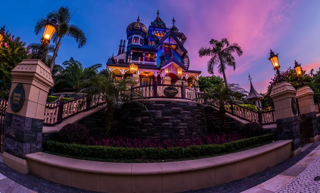 The illuminated Victorian-style Mystic Manor mansion with domes and spires glowing against a purple and pink sunset sky, featured in a Hong Kong Disneyland Guide.