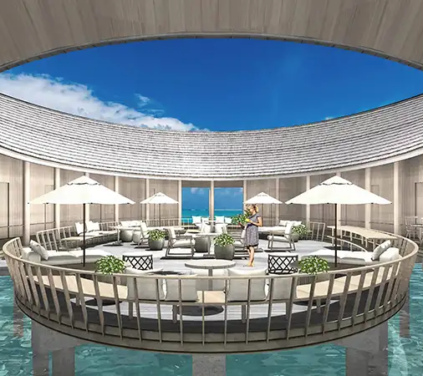 Maldives Island Tour - An interior architectural photograph looking into the central circular open-air courtyard of an overwater spa lounge built on stilts over clear turquoise waters in the Maldives, featuring white sun umbrellas, lounge seating, wooden timber pillars, and a massive circular roof opening revealing a bright blue sky.