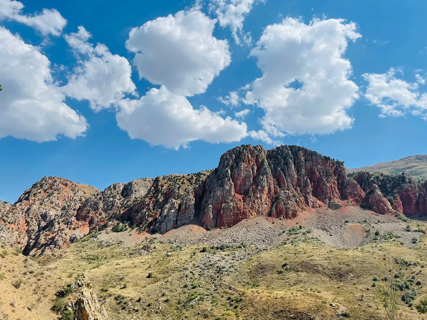 A wide, low-angle landscape photograph showing towering red and ochre limestone cliffs rising above arid, grassy hillsides and scree fields under a vibrant blue summer sky with fluffy white cumulus clouds near Noravank, Armenia.