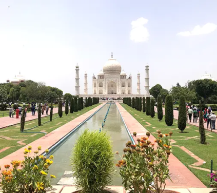 India and Maldives Tour - A wide landscape photograph looking down the central reflecting pool toward the white marble Taj Mahal mausoleum, flanked by cypress trees, green lawns, and garden pathways with visitors walking under a bright, sunny sky in Agra, India.