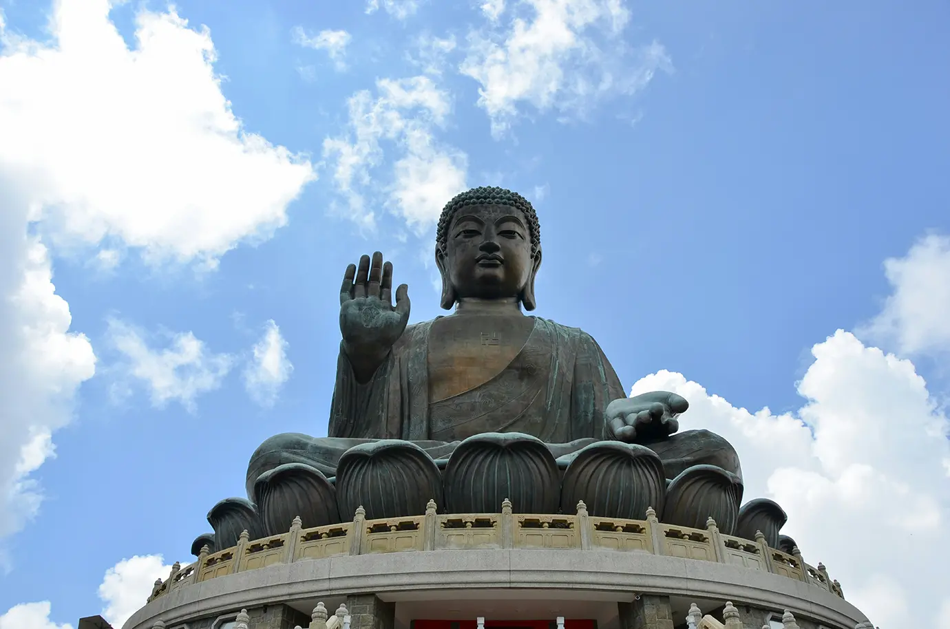 A low-angle view of the large bronze Tian Tan Buddha seated on a lotus pedestal against a bright blue sky with white clouds, showcasing cultural landmarks for Things to Do in Hong Kong. 