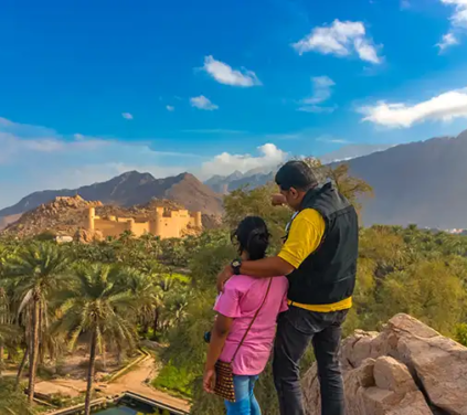 Oman and Maldives Tour - A high-angle outdoor travel photograph of a man and woman standing on a rocky outcrop, pointing toward the mud-brick Nakhal Fort nestled within a lush green date palm grove and backed by the Hajar Mountains in Nakhal, Oman.