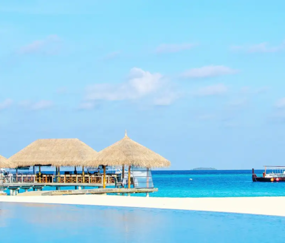 A wide landscape photograph showing the calm, transparent blue waters of a shallow ocean lagoon in the foreground, bordering a white sand beach, leaning coconut palm, thatched-roof resort pavilions, and a traditional dhoni boat in South Malé Atoll, Maldives.