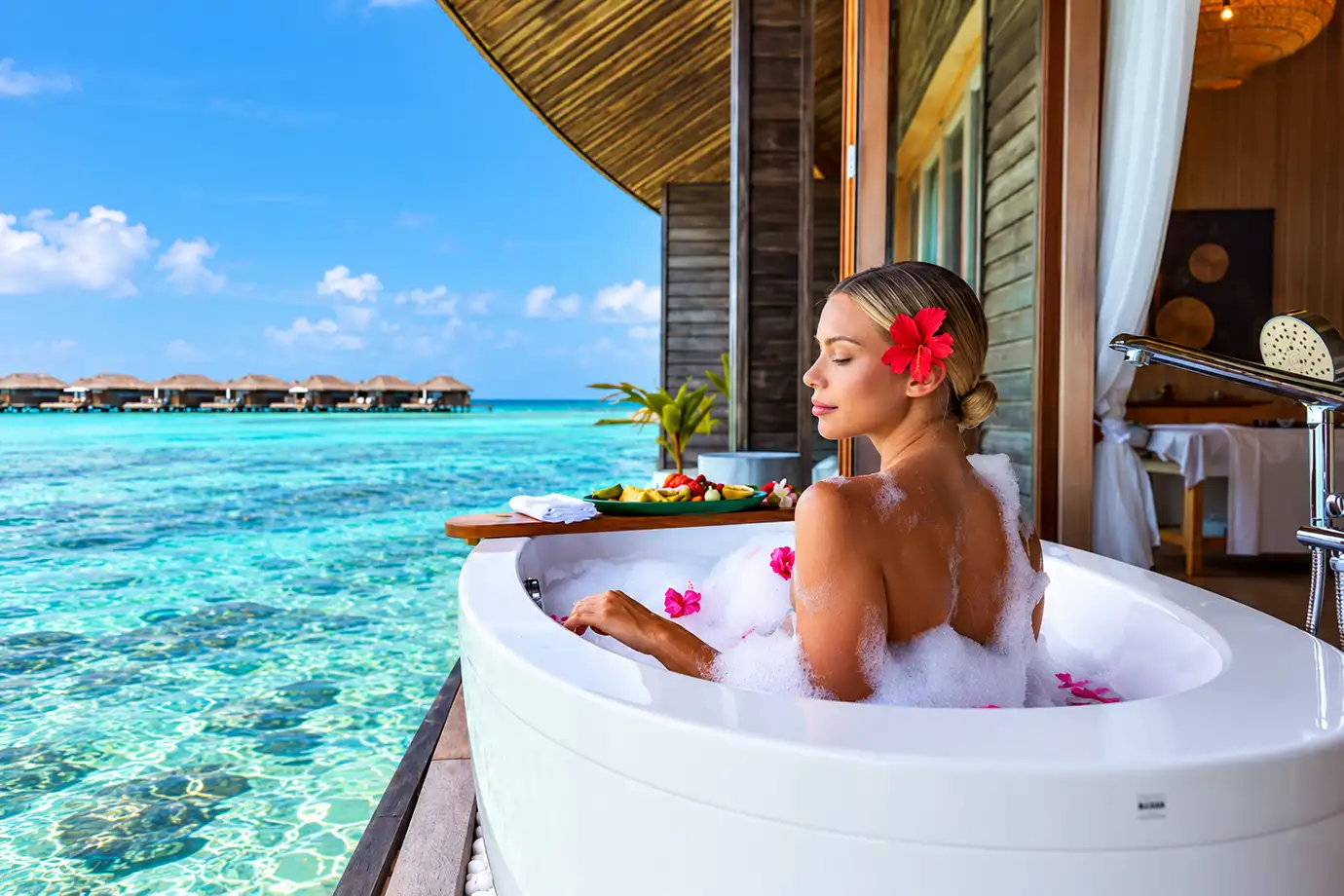 A eye-level travel photograph of a woman with a red hibiscus flower tucked behind her ear relaxing in a bubble bath inside a freestanding white bathtub on a private wooden deck, overlooking turquoise lagoon waters and overwater villas at Kagi Maldives Resort & Spa.