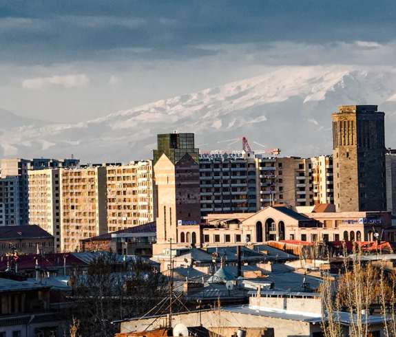 A wide cityscape photograph taken from an elevated perspective looking across the rooftops and modern architecture of Yerevan, Armenia, toward the snow-capped peak of Mount Ararat under a dramatic, cloudy sky.